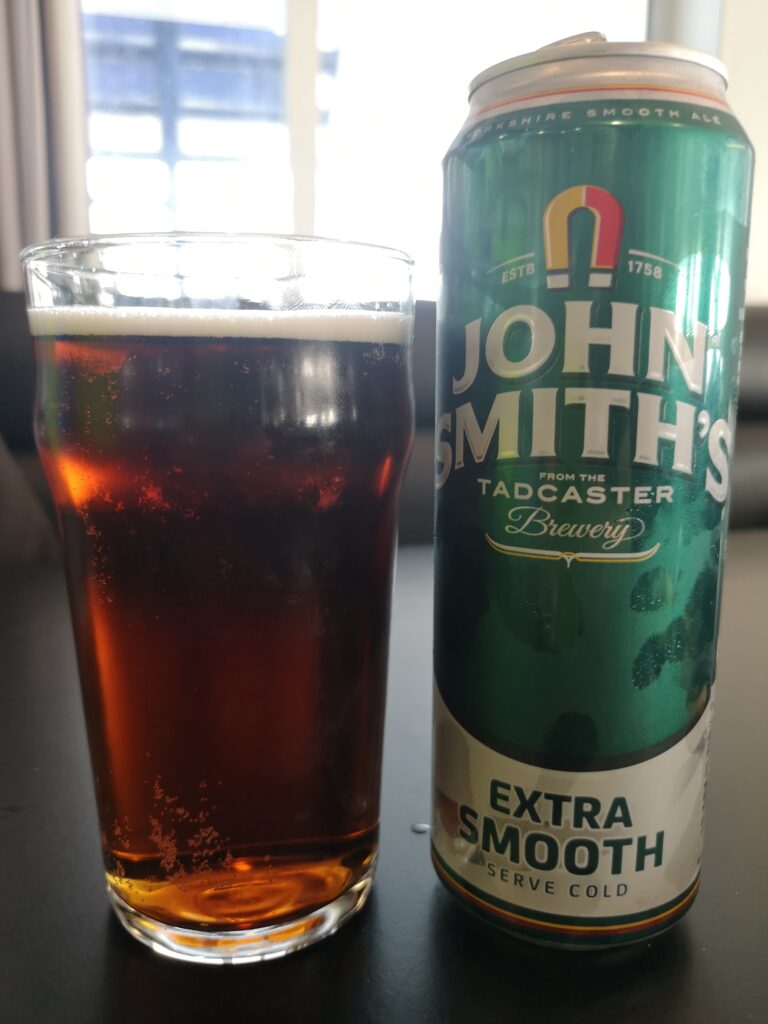 Vor uns auf einem Tisch steht ein Glas gefällt mit braunem Ale und daneben eine britishe Bierbüchste mit der Aufschrift "John Smiths Extra smooth"