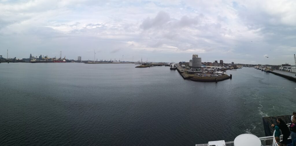 Blick von der Hafenausfahrt zurück auf den Hafen von Ijumieden. Der linke Bereich ist mit Industrianlagen und allerlei Türmen und Schornschteinen gefüllt, davor das Hafenbecken. Auf der rechten Seite sieht man das Hafenbecken, von wo unsere Fähre gerade abgelegt hat 