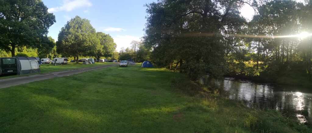 Sonnenaufgang am Zeltplatz Cobleland bei Aberfoyle.

Rechts im Bild sehen wir einen Fluss und das Ufer ist mit Bäumen gesäumt, durch die die Sonne durch Blätterlücken durchscheint.
Vor uns ist ein Wiesenstreifen, auf dem weiter hinten noch ein Zelt und ein Auto steht. neben dem Streifen ist eine aspahltierte Fahrspur und links neben der, wieder eine Wise, auf der Camper und zelte stehen