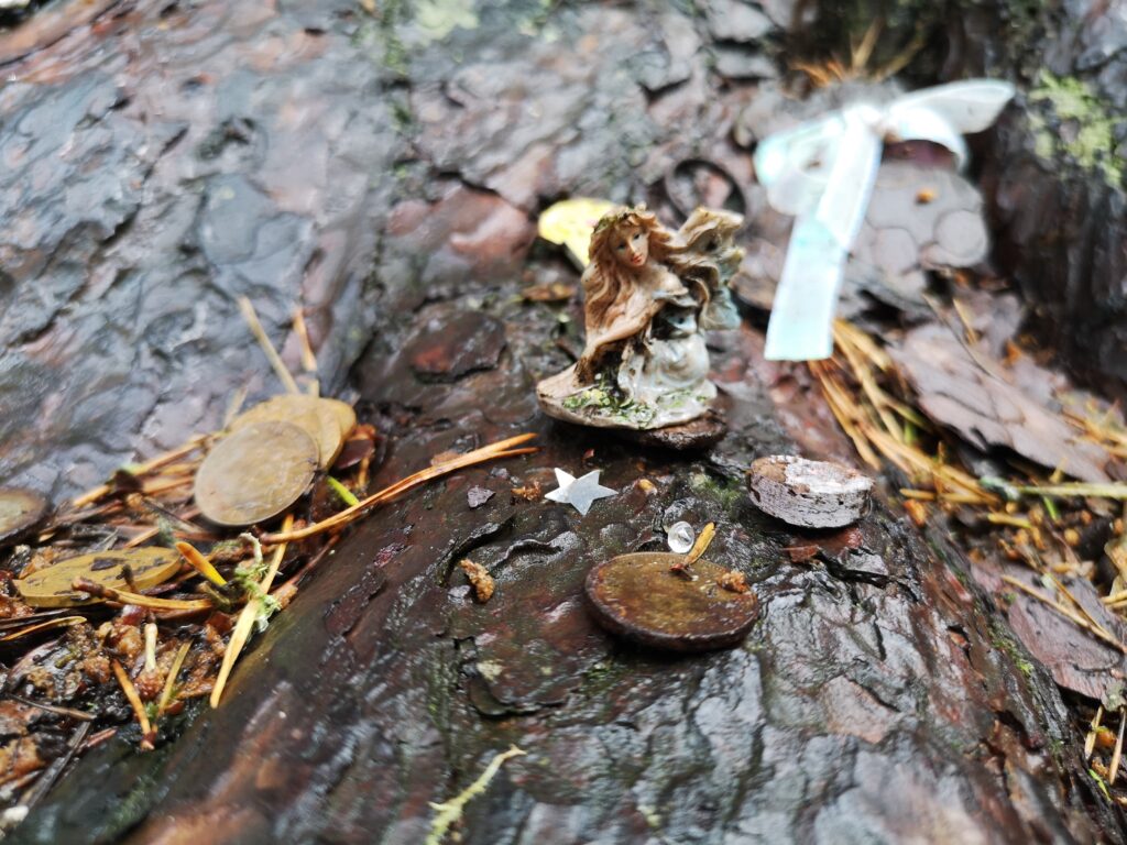 Auf einem Teil der Wurzel am Boden ist eine ca. 3 cm hohe kleine Feeenskulptur. Rings herum liegen Münzen um den Baum und auch bei der kleinen Fee