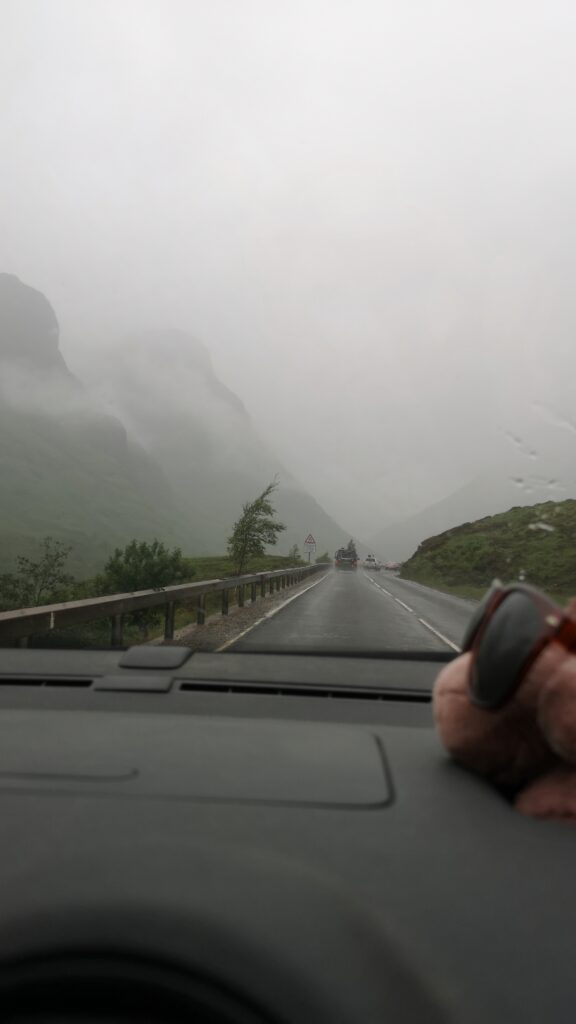Wir fahren durch das bekannte Tal Glen Coe in Schottland. Es regnet und von den hohen Bergen links und rechts der Straße, ist so gut wie nichts zu sehen, weil die Wolken bis fast auf Straßenniveau tief hängen. Seitlich sieht man die Konturen von steilen Berghängen