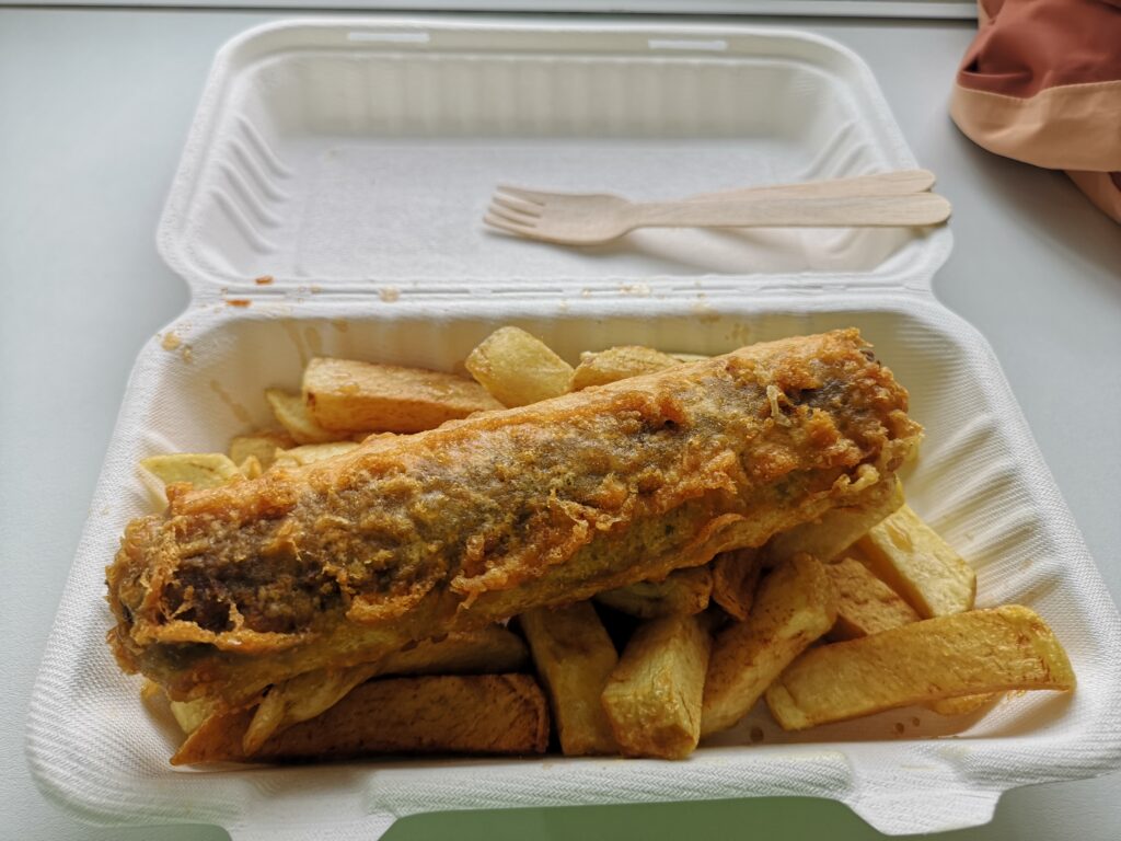 Vor uns liegt eine Portion Haggis udn Chips.
Der Haggis ist in einer kleinen Wurstform und außen mit Bierteig umhüllt und frittiert worden. 
Er liegt oben auf einer Portion schottischer Pommes, die in UK Chips genannt werden.
Beides liegt auf einem Fensterbrett in einer to go Pappverpackung in der Chippie Bude