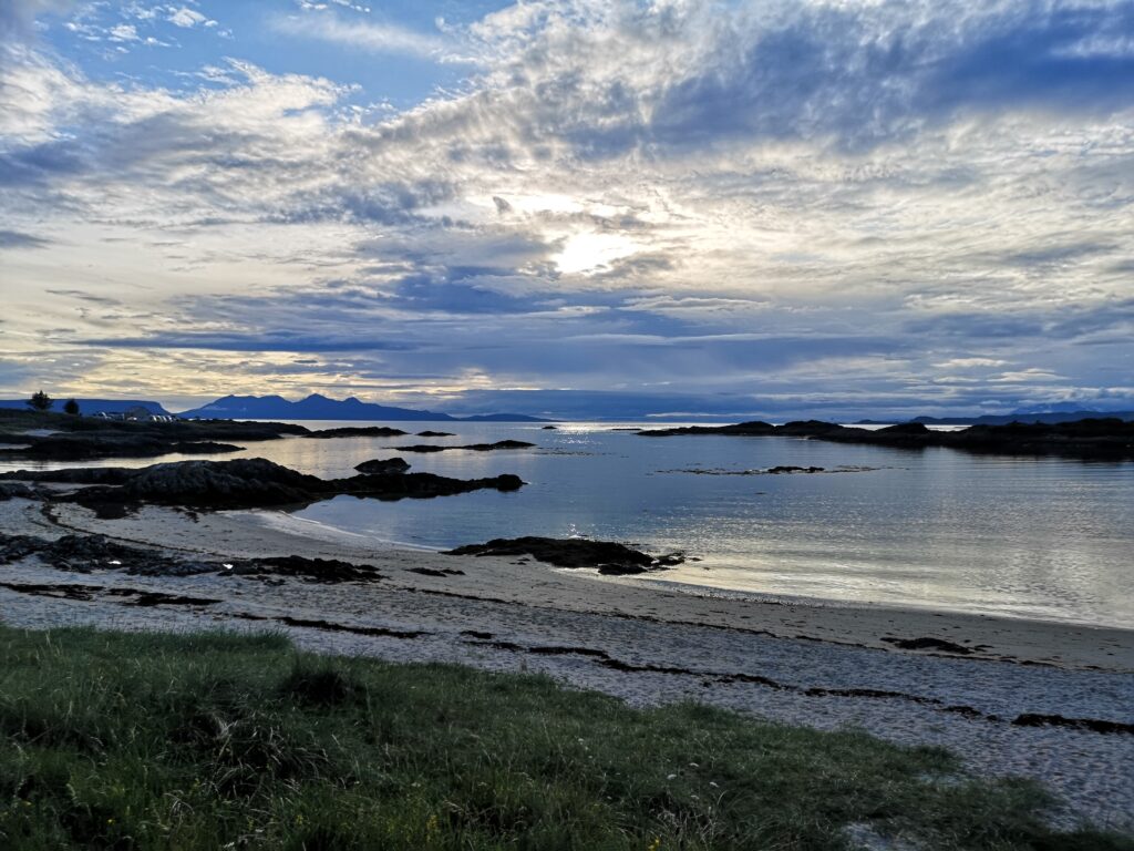 Wir stehen am Ufer am Traigh Golf Course und schauen Richtung Westen.
Unter uns ist weißer Sandstand.
 Die Sonne ist durch Schleierwolken verdekt und beleuchtet die Umgebung indirekt und man sieht leichte Wolken im Meer wiederspiegeln