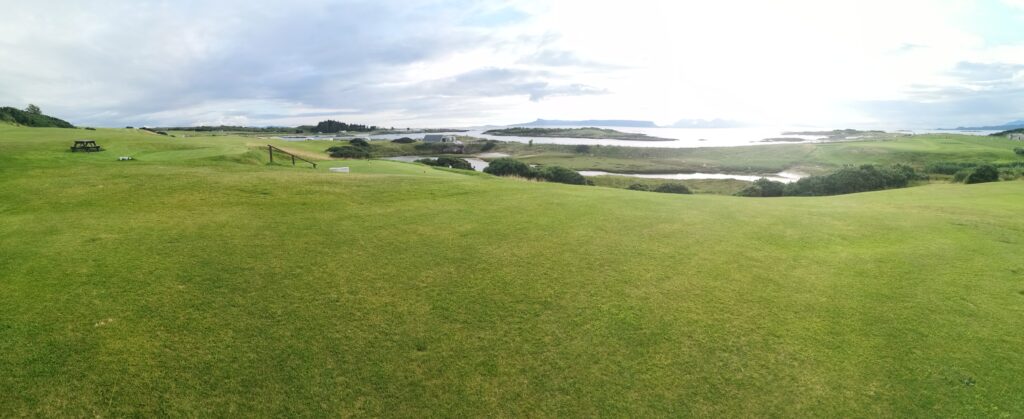 Blick von oben auf dem Golfplatz in Richtung Amerika. Vor uns breitet sich die Wiese des Golfplatzes bis zum Ufer aus. Dieser besteht aus weißem Sand und das Meer schimmert Türkisfarben zurück.
Im Meer sieht man in einiger Entfernung noch ein paar Inseln 