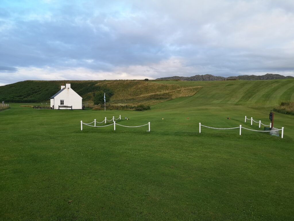 Wir stehen auf einer sehr grünen Wiese und haben einen kleinen weißen Zaun in halber Entfernung stehen, der aus weißen Pfosten udn zischen diesen einem Strick besteht.
Am linken mittleren Hintergrund steht das Clubhaus des Traigh Golf Course und dahiunter erheben sich grün bewachsene Dünen