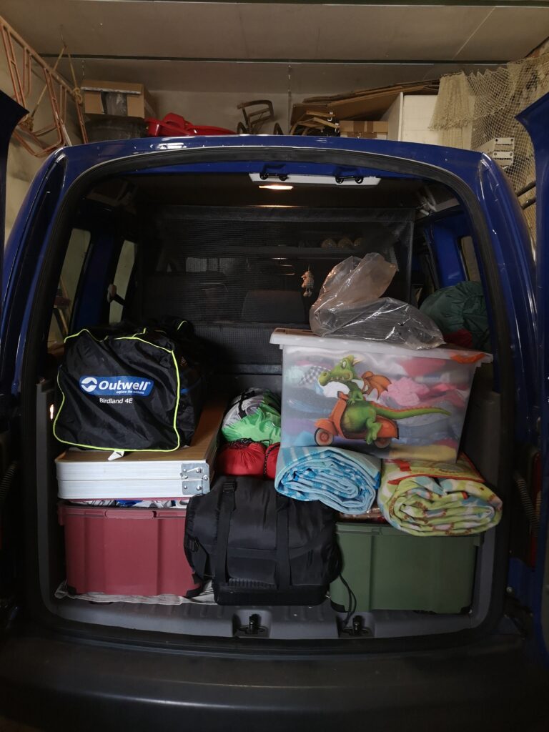 Ein VW Caddy dessen hintere Türen offen sind und der Laderaum zur Hälfte beladen ist mit Zelt, Klappkisten, Schlafsäcke, Decken und Proviantkisten