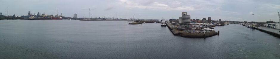 Blick von der Hafenausfahrt zurück auf den Hafen von Ijumieden. Der linke Bereich ist mit Industrianlagen und allerlei Türmen und Schornschteinen gefüllt, davor das Hafenbecken. Auf der rechten Seite sieht man das Hafenbecken, von wo unsere Fähre gerade abgelegt hat
