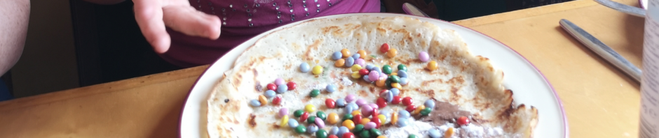 wir sehen auf einen Teller auf einem Pannekukenboot in Rotterdam. Auf diesem Teller liegt ein Eierkuchen, der mit Smarties, Nussnugatcreme und Puderzucker belegt ist und gleich gegessen wird