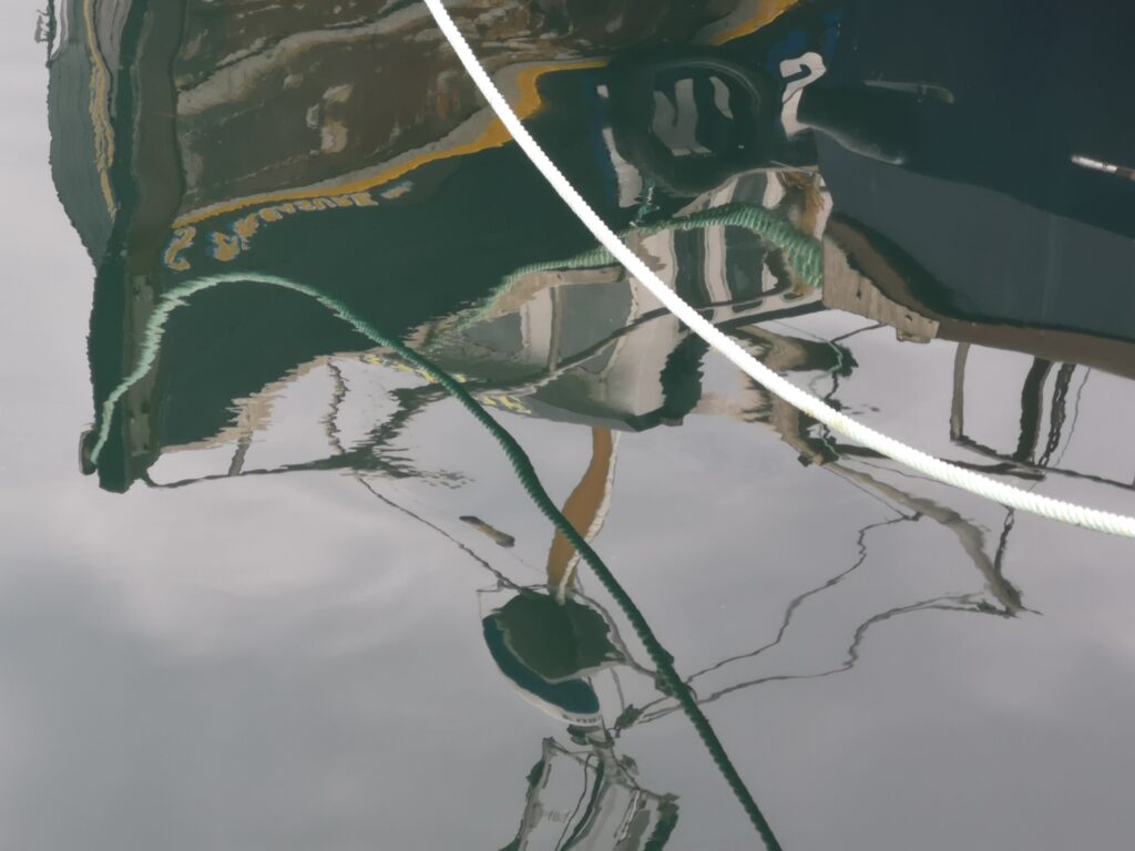 Wir blicken auf eine Spiegelung im Hafen von Mallaig, wo man die Konturen eines Fischerbootes sehen kann mit seinen Aufbauten. Quer durch das Bild geht einen über dem Wasser hängende Halteleine eines Bootes