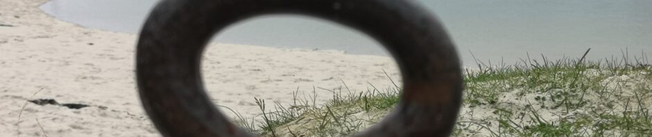 Blick durch einen Eisenring am Ufer der White Sands of Morar, auf eben diesen weißen Sandstrand. Rechts im Bild sieht man eine Flussmündung, die ins Meer geht und auf der linken Bildseite, den weißen Sandstrand, der aus dem Film "lokal Hero" bekannt ist